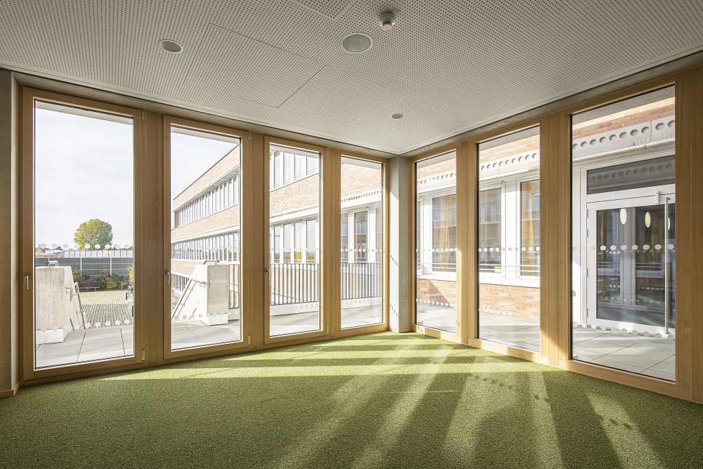 Heller Innenraum mit großen Holzrahmen-Fenstern, die auf einen Innenhof und ein modernes Schulgebäude blicken. Teppichboden in Grün und perforierte Deckenpaneele.