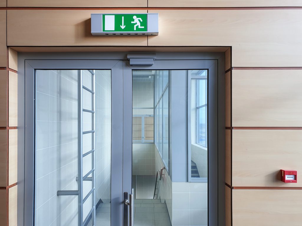 Eine gläserne Notausgangstür in einem Gebäude. Darüber hängt das entsprechende grüne Schild.