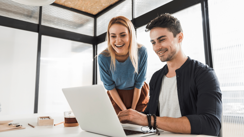 Ein junger Mann und eine junge Frau stehen zusammen an einem Schreibtisch in einem modernen, hellen Büro mit Glaswänden. Beide schauen lächelnd auf einen geöffneten Laptop, der auf dem Tisch steht. Der Mann sitzt und tippt, während die Frau sich von hinten leicht über ihn beugt und freundlich mitlacht. Auf dem Tisch liegen ein Glas Tee, einige Stifte und ein kleines Kästchen mit Büroklammern. Die Atmosphäre wirkt freundlich, konzentriert und kollegial.