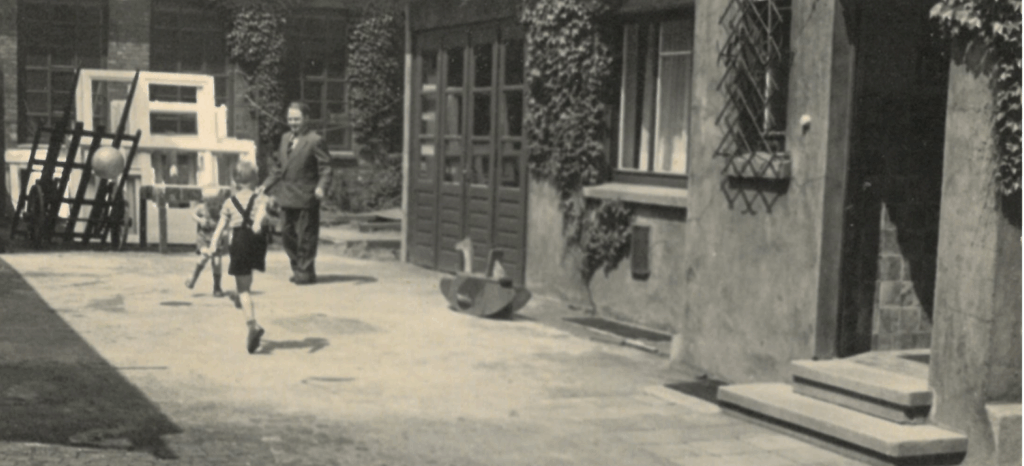 Zwei Jungen rennen mit Holzstöcken durch einen Hof, während ein Mann ihnen lächelnd zusieht. Im Hintergrund sind große Fensterrahmen und Werkstattgegenstände zu erkennen. Die Szene zeigt den Alltag im Ruhrgebiet um 1960 – geprägt von Arbeit und gleichzeitig von familiärem Leben mitten im Handwerksbetrieb.