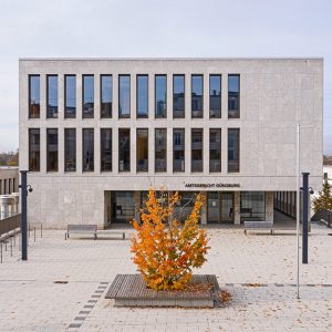 Das Amtsgericht Günzburg, ein modernes Gebäude mit einer Fassade aus hellem Stein und vertikalen Fensterreihen, dominiert einen gepflasterten Platz. Im Vordergrund steht ein Baum mit leuchtend orangefarbenem Herbstlaub.