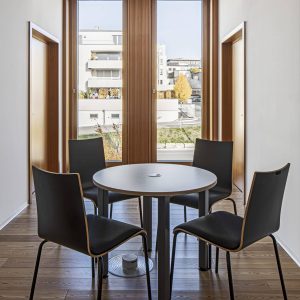 Ein minimalistischer Besprechungsraum mit vier schwarzen Stühlen um einen runden Tisch, platziert auf einem eleganten Holzboden. Im Hintergrund befinden sich hohe Holzrahmenfenster mit perforierter Verkleidung.