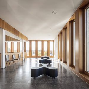 Ein heller, moderner Innenraum mit großen Holzrahmenfenstern, die eine weite Aussicht und reichlich Tageslicht bieten. Der Boden besteht aus polierten dunklen Steinfliesen, die einen eleganten Kontrast zu den warmen Holzwänden schaffen.