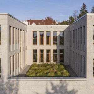 Nahaufnahme des Innenhofs des Amtsgerichts Günzburg, eingerahmt von der hellen Kalksteinfassade des Gebäudes mit symmetrisch angeordneten Fenstern. Im Zentrum des Innenhofs befindet sich eine begrünte Fläche mit niedrigen Pflanzen und jungen Bäumen.