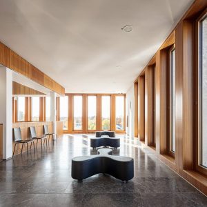 Ein heller Innenraum mit großen Holzrahmenfenstern, die viel Tageslicht hereinlassen, und einer modernen Einrichtung mit schwarzen Designer-Sitzmöbeln sowie Stühlen entlang der Wand. Der Steinboden und die klaren Linien schaffen eine elegante und einladende Atmosphäre.