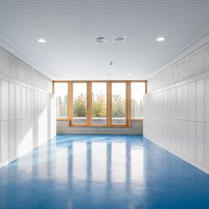 Heller, moderner Flur mit glänzendem blauem Boden, flankiert von weißen Schließfächern auf beiden Seiten. Große Fenster mit Holzrahmen lassen Tageslicht herein und bieten einen Blick auf eine grüne Außenanlage.