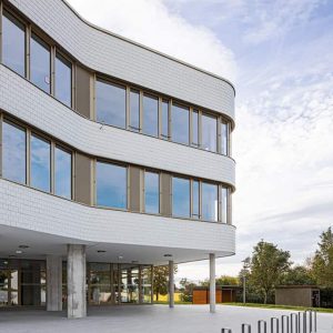 Modernes Gebäude mit geschwungener, weiß gekachelter Fassade und großen Fenstern. Der Eingangsbereich ist von schlanken Pfeilern gestützt, umgeben von Bäumen im Hintergrund.