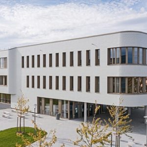 Modernes, weiß verkleidetes Gebäude mit abgerundeten Ecken und großen Fenstern. Es ist dreigeschossig und von einer grünen Rasenfläche mit jungen Bäumen umgeben. Der Eingangsbereich ist überdacht, und der Platz davor ist gepflastert.