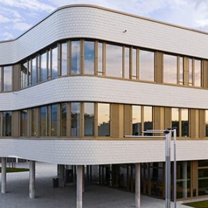 Modernes Gebäude mit geschwungenen Fassaden und großflächigen Fenstern, die den Abendhimmel reflektieren. Die Fassade ist mit hellen Kacheln verkleidet, der Eingangsbereich liegt auf Pfeilern. Umgeben von einer Grünfläche und einem gepflasterten Vorplatz.