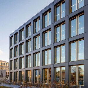 Moderne Bürogebäude-Fassade mit großen, rechteckigen Fenstern in einem dunklen, geradlinigen Rahmen. Die Fenster reflektieren den blauen Himmel und umliegende Gebäude, während das Abendlicht die Struktur warm beleuchtet.