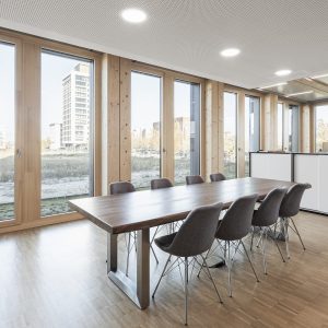 Moderner Besprechungsraum mit einem großen Holztisch und acht gepolsterten Stühlen. Große Holzrahmenfenster lassen viel Tageslicht herein und bieten Blick auf umliegende Gebäude und eine Grünfläche.