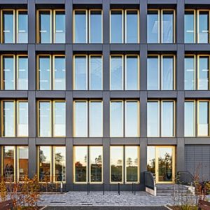 Frontansicht eines modernen Bürogebäudes mit symmetrischer Fensteranordnung, rechteckigen Glasflächen und dunklen Rahmen. Die Fassade reflektiert die Umgebung und das weiche Abendlicht.