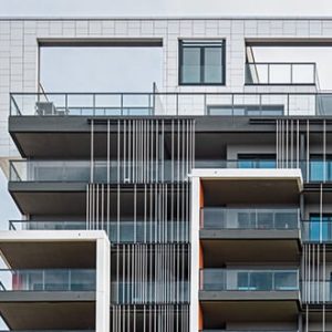 Detailansicht der oberen Etagen eines modernen Wohngebäudes mit großzügigen Balkonen, bodentiefen Fenstern und auffälligen vertikalen Metallstreben als Gestaltungselemente. Die Dachpartie umfasst offene Rahmenstrukturen und Glasgeländer.