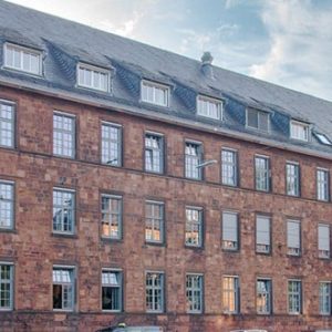 Breites historisches Gebäude aus rotem Sandstein mit vielen Fenstern und einem geneigten Schieferdach. Typische Architektur des frühen 20. Jahrhunderts, ergänzt durch Gauben und schlichte Details.