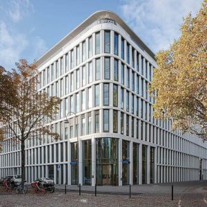 Moderne Bürogebäude-Fassade mit abgerundeten Ecken und vertikalen Fensterstreben, umgeben von herbstlichen Bäumen. Im Vordergrund sind Fahrräder und Pflastersteine sichtbar.