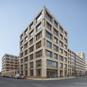 Modernes Bürogebäude aus beigefarbenem Naturstein und großen Holzrahmenfenstern, das klare Linien und geometrisches Design vereint. Im Erdgeschoss befinden sich Eingänge und eine Tiefgarageneinfahrt, während Autos und Fahrräder entlang der Straße geparkt sind.