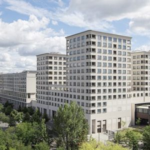 Panorama eines modernen Hotelkomplexes mit heller Fassade und symmetrischen Fenstern, umgeben von Bäumen, Parkanlagen und angrenzenden Stadtgebäuden.