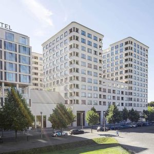 Moderner Hotelkomplex mit einer Fassade aus hellem Putz und großen, symmetrisch angeordneten Fenstern. Die Baukörper mit abgerundeten Ecken sind terrassenartig angeordnet und von einer begrünten Umgebung mit Bäumen und parkenden Autos umgeben.