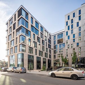 Ein modernes Gebäude mit heller Steinfassade, großen Glasfenstern und einer klaren, geometrischen Architektur, das sich über mehrere Etagen erstreckt. Im Vordergrund parken Autos entlang der Straße, während Bäume und blauer Himmel eine freundliche Atmosphäre schaffen.