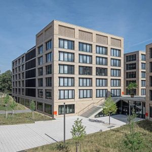 Helles Bürogebäude aus Klinkerstein mit großen, regelmäßig angeordneten Fenstern und moderner Architektur. Im Vordergrund befindet sich ein weitläufiger, gepflasterter Vorplatz mit Treppen, jungen Bäumen und einer Grünfläche.