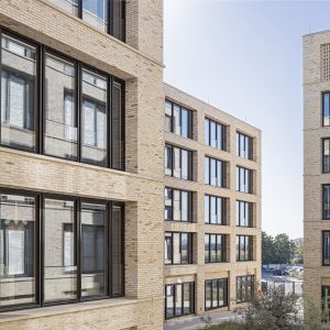 Mehrere moderne Bürogebäude mit Ziegelfassade und großen schwarzen Fenstern, die in einer urbanen Umgebung angeordnet sind. Im Vordergrund und Hintergrund sind helle Fassaden und Fensterfronten zu sehen, umgeben von einem schmalen Innenhof und vereinzelter Begrünung.