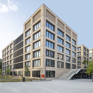 Modernes, sechsgeschossiges Gebäude mit heller Klinkerfassade und großen, symmetrischen Fensterfronten, eingebettet in eine urban gestaltete Umgebung mit gepflasterten Wegen und jungen Bäumen. Im Vordergrund führen Treppen zu einem Eingangsbereich, während der blaue Himmel das klare Design der Architektur betont.