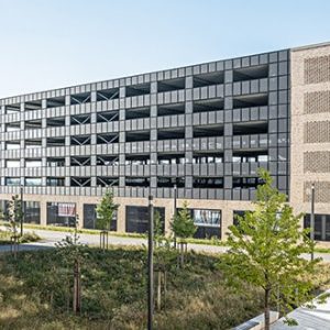 Moderne Architektur mit einem mehrstöckigen Parkhaus aus dunklen Fassadenelementen und seitlichen Backsteinflächen. Im Vordergrund befinden sich begrünte Außenanlagen mit jungen Bäumen und gepflasterten Wegen.